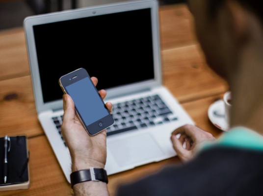 Close up of a person at a desk holding a phone in front of a laptop